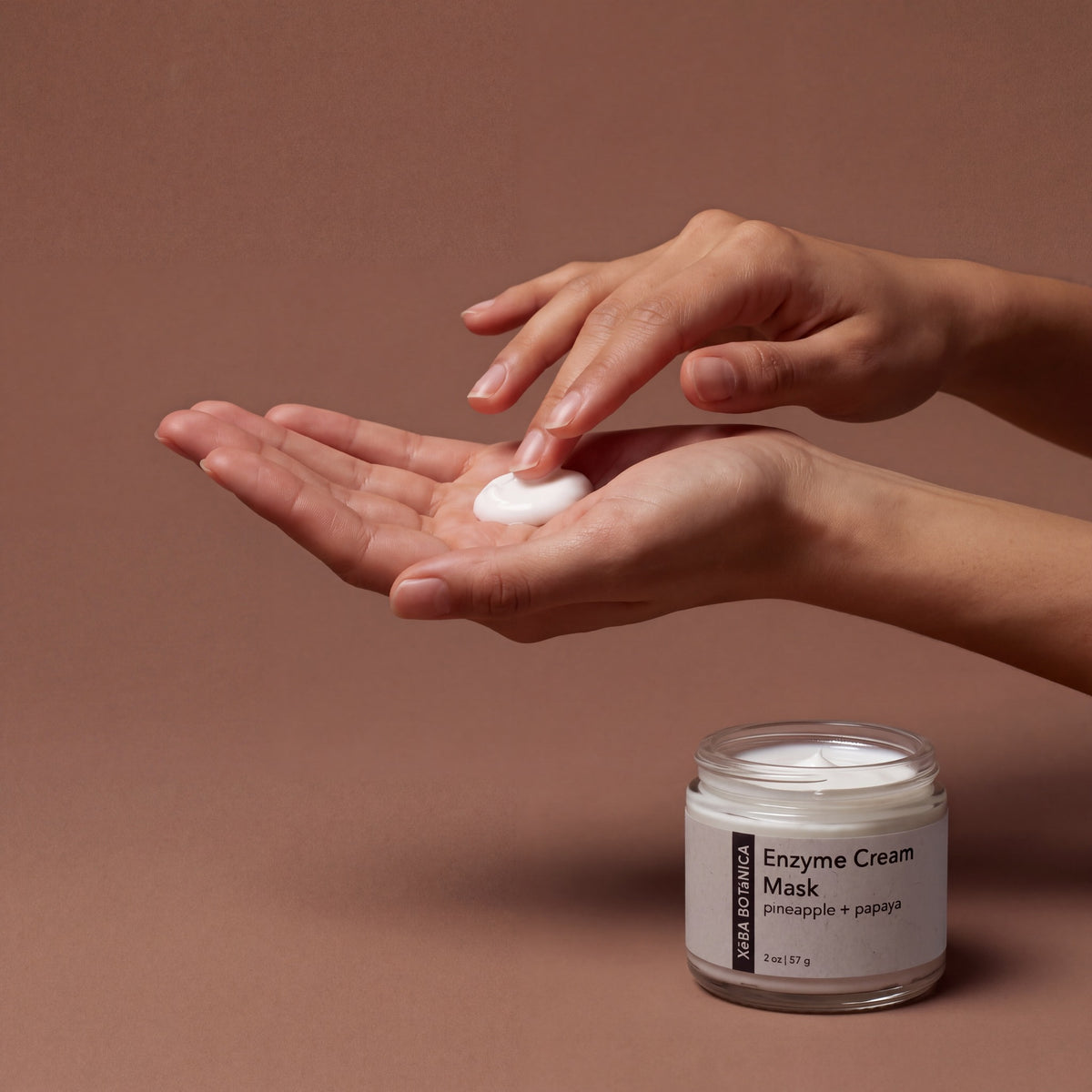 Hands holding a small dab of Enzyme Cream Mask beside the open jar