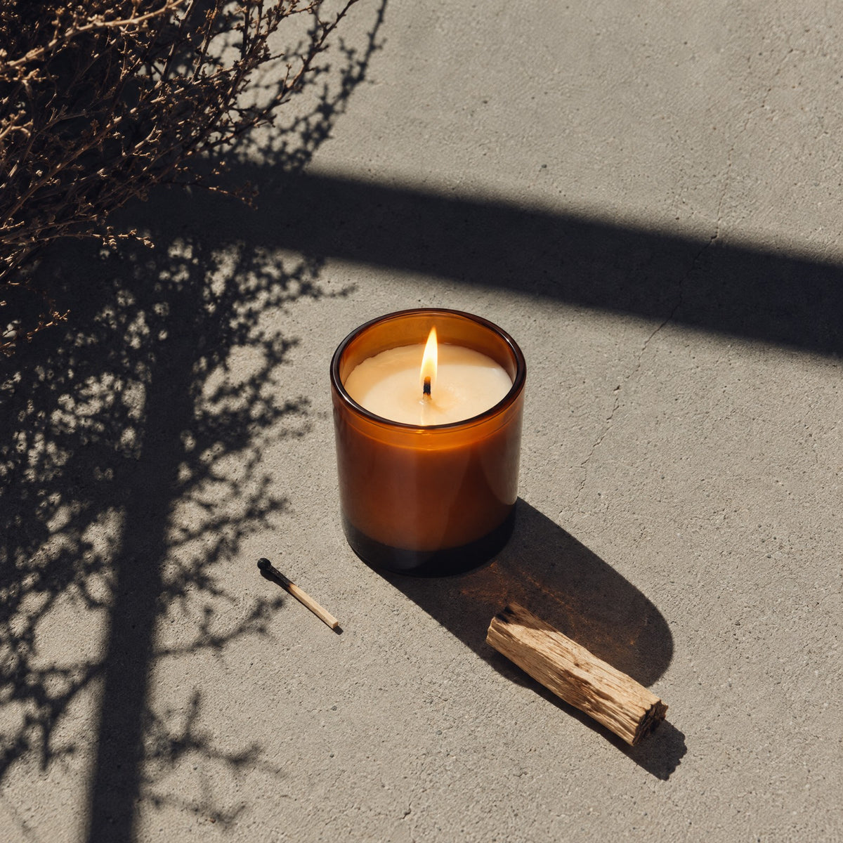 Lit Amber Glass Candle on sunlit concrete beside a palo santo stick