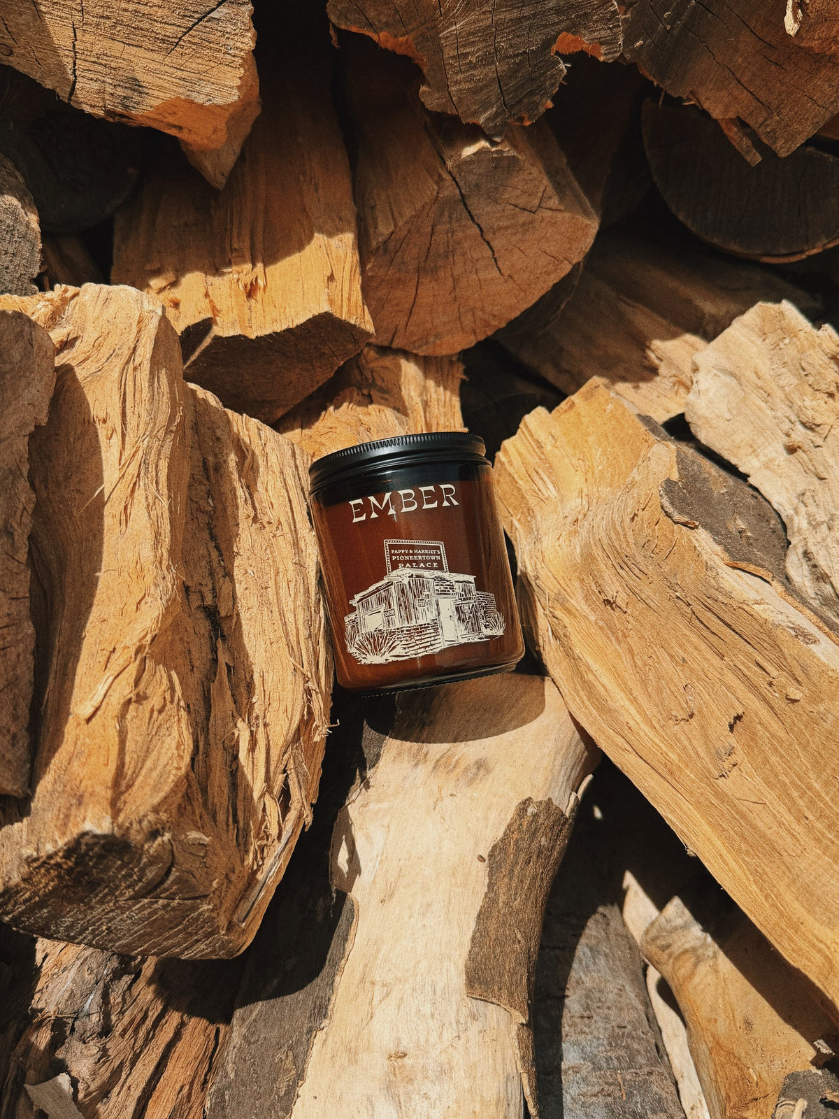 Ember candle in an amber glass jar resting on a stack of split firewood in desert sun