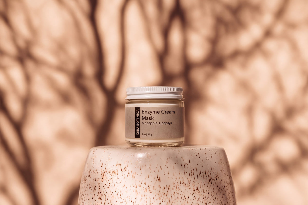 Enzyme Cream Mask jar on a speckled ceramic pedestal against a warm sand wall